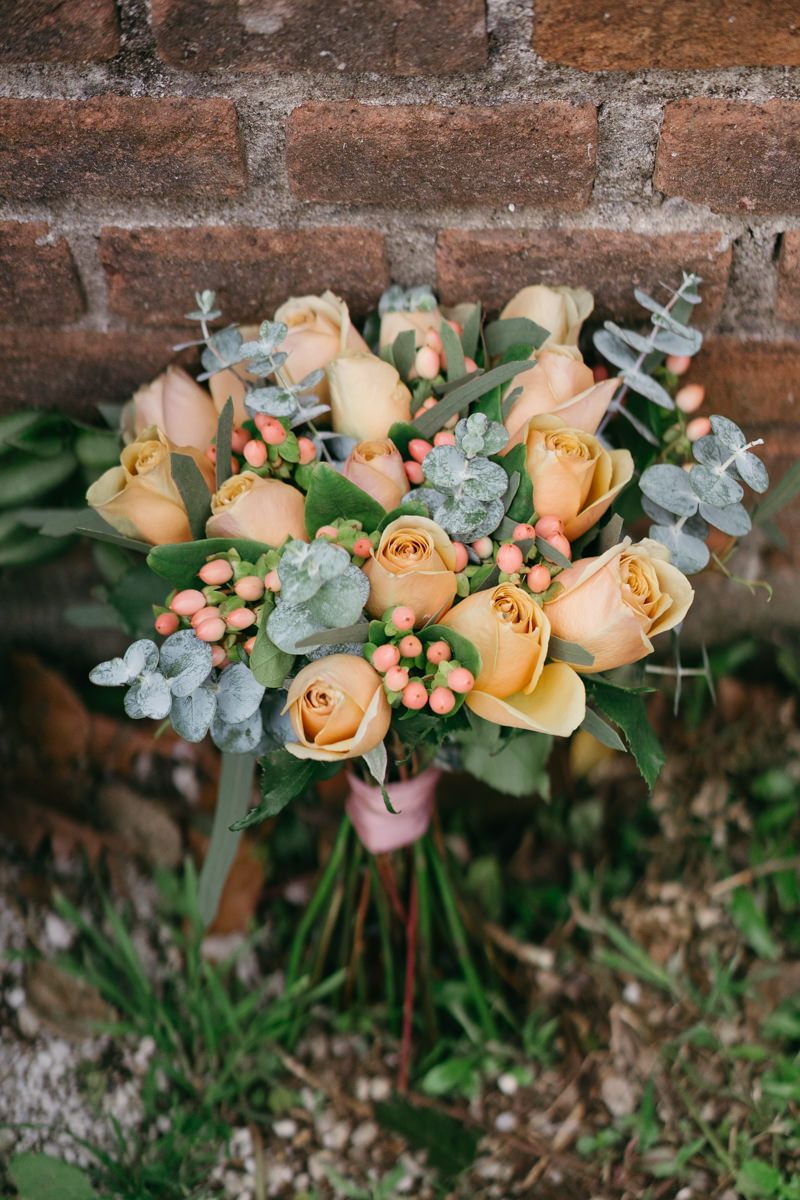 Close-up of peachy-orange roses with eucalyptus leaves arranged against weathered brick background.