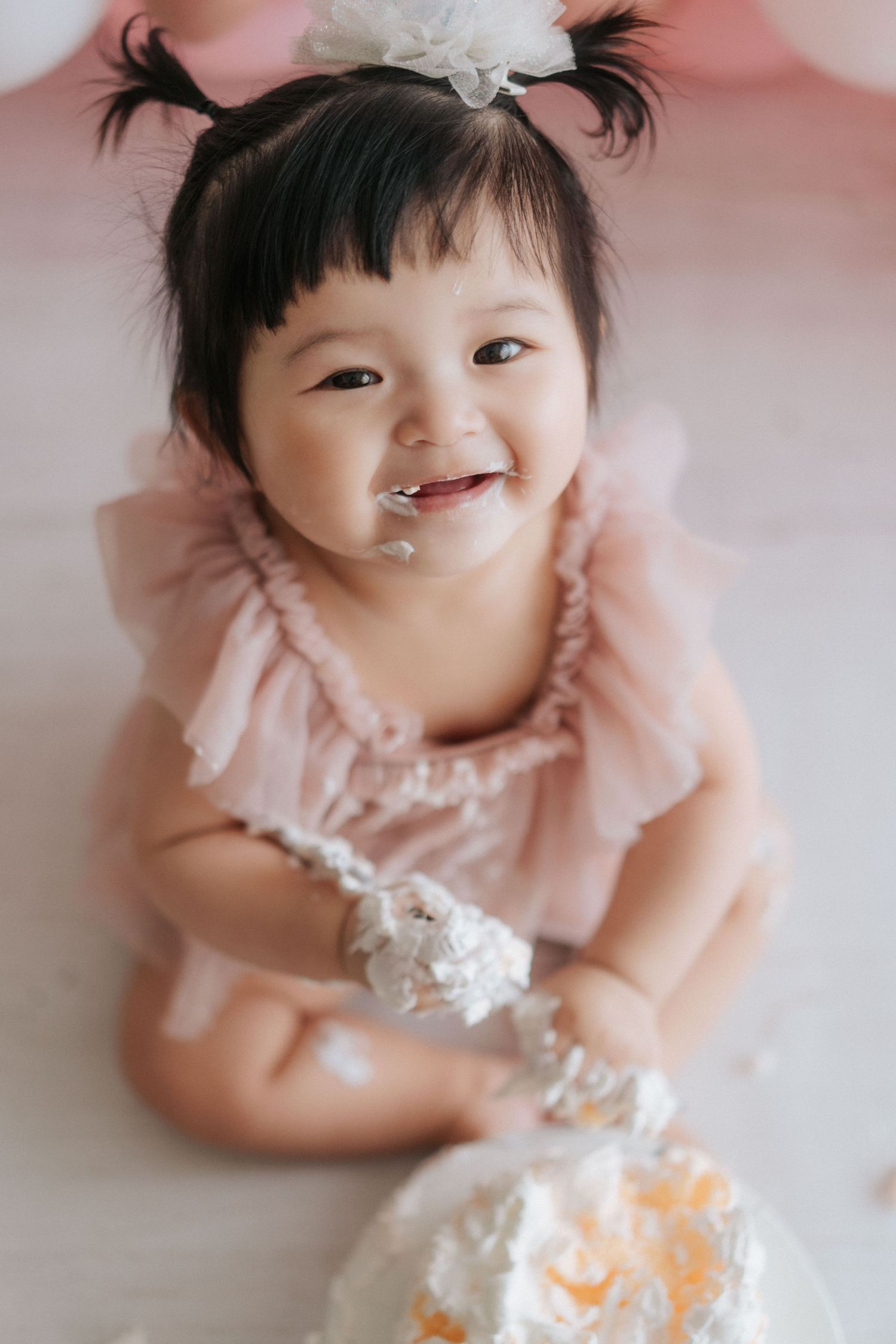 Baby in pink ruffled dress smiles joyfully while playing with cake during first birthday cake smash photo session.