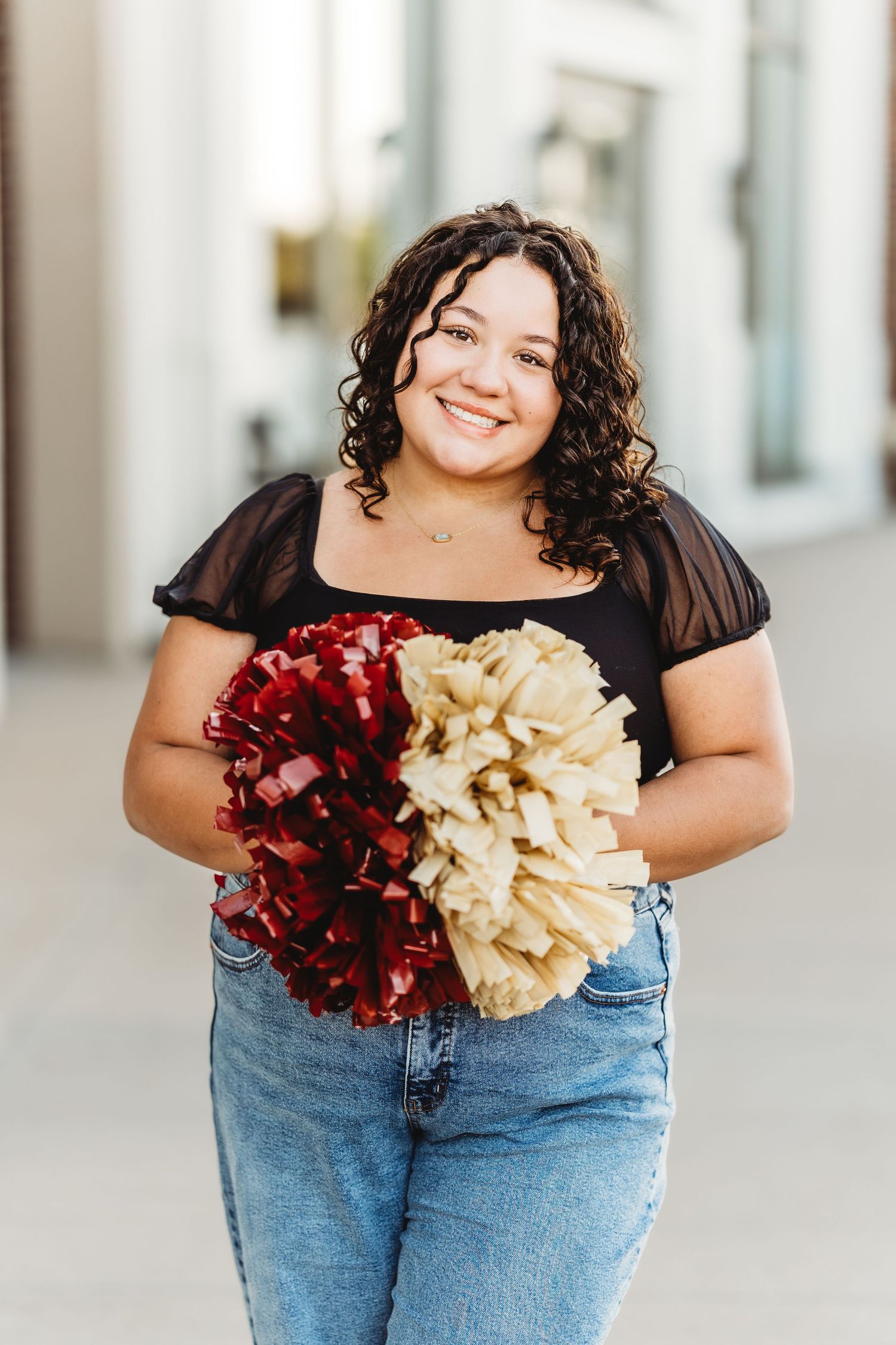 A person in jeans and black top holds red and gold cheerleading pom poms in front of a light colored wall.