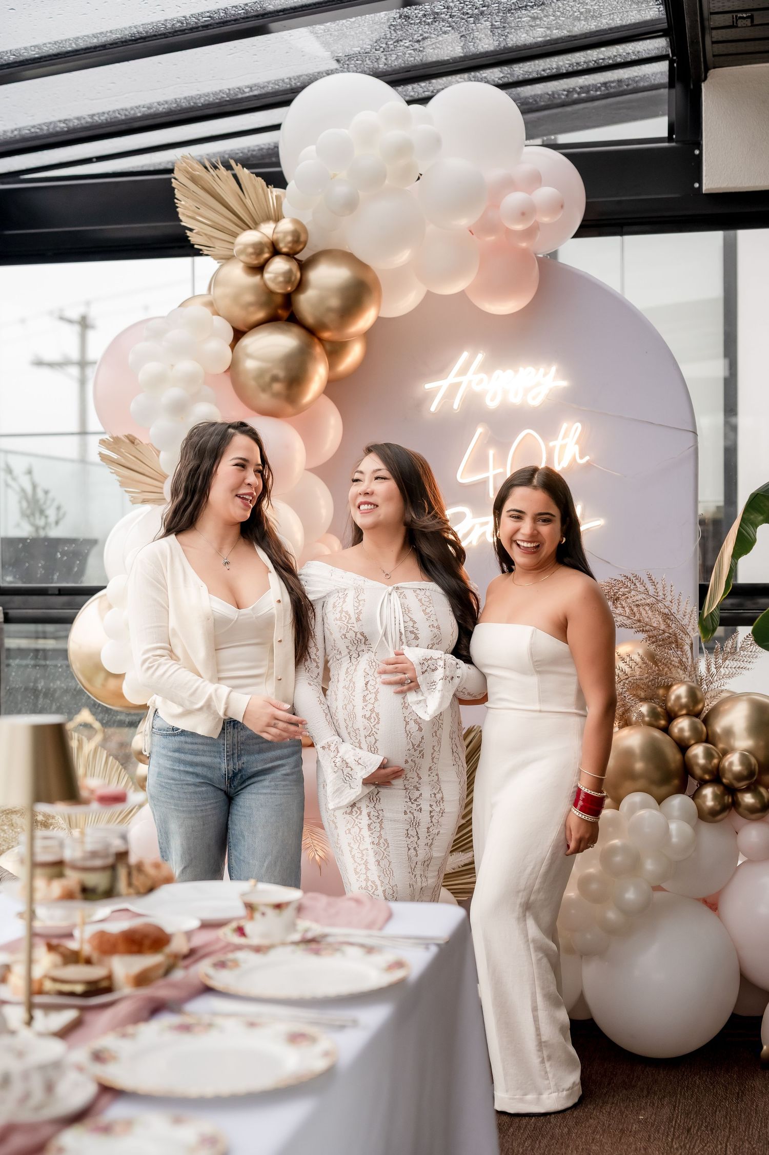 Elegant birthday celebration setup with rose gold and white balloon arch, neon sign, and woman in sequined dress.