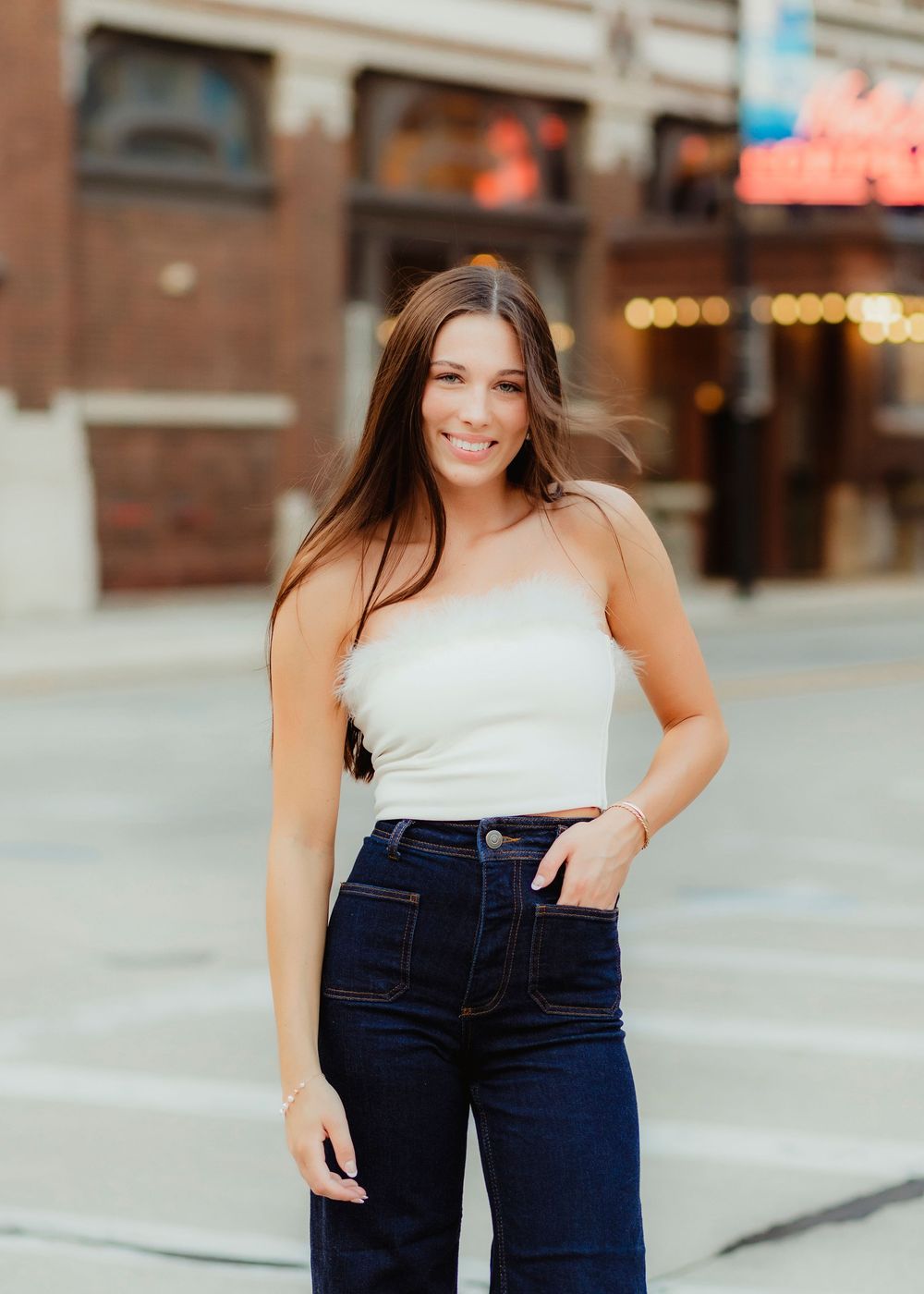 A person in a white top and dark jeans poses in an urban setting with brick buildings.