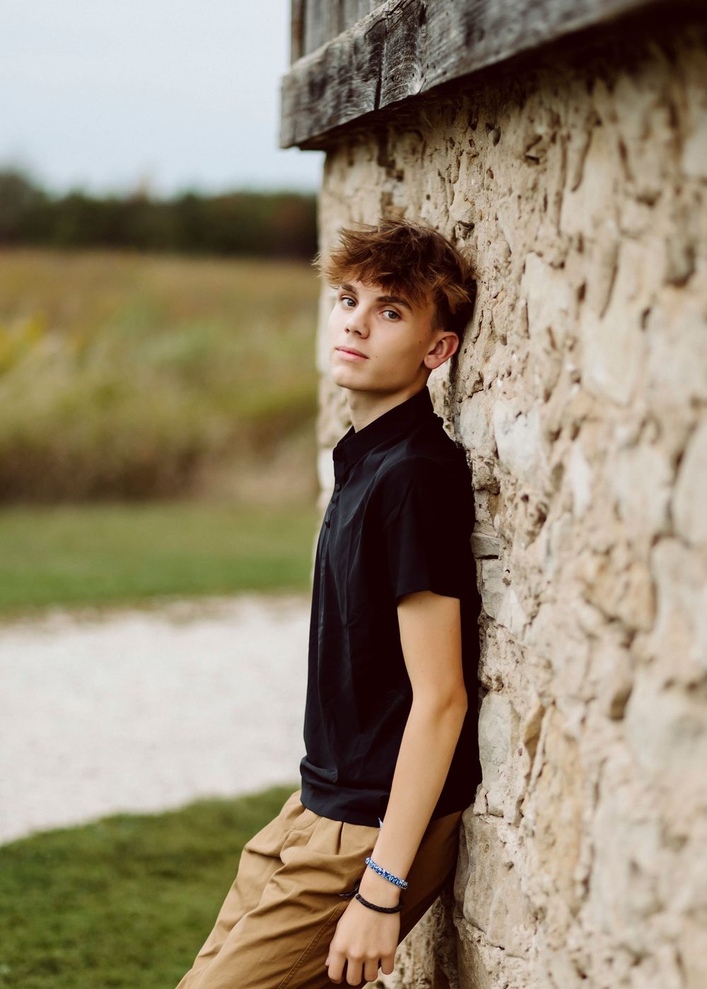 Outdoor portrait against rustic stone wall wearing black polo shirt.