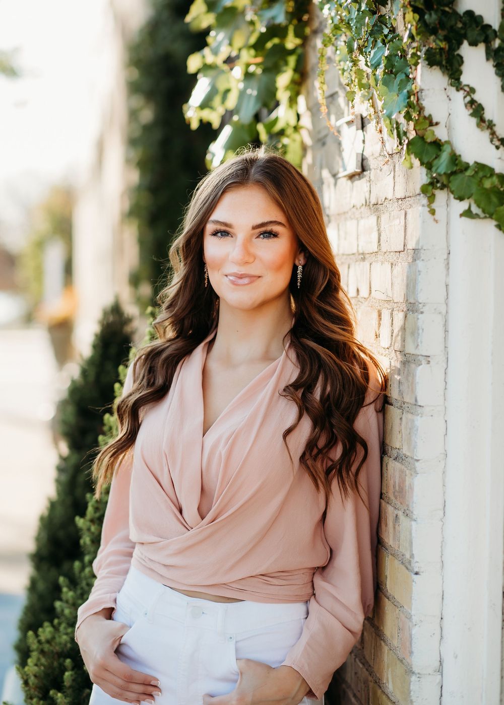 Fashion portrait featuring soft pink wrap blouse and white pants against brick wall with greenery.