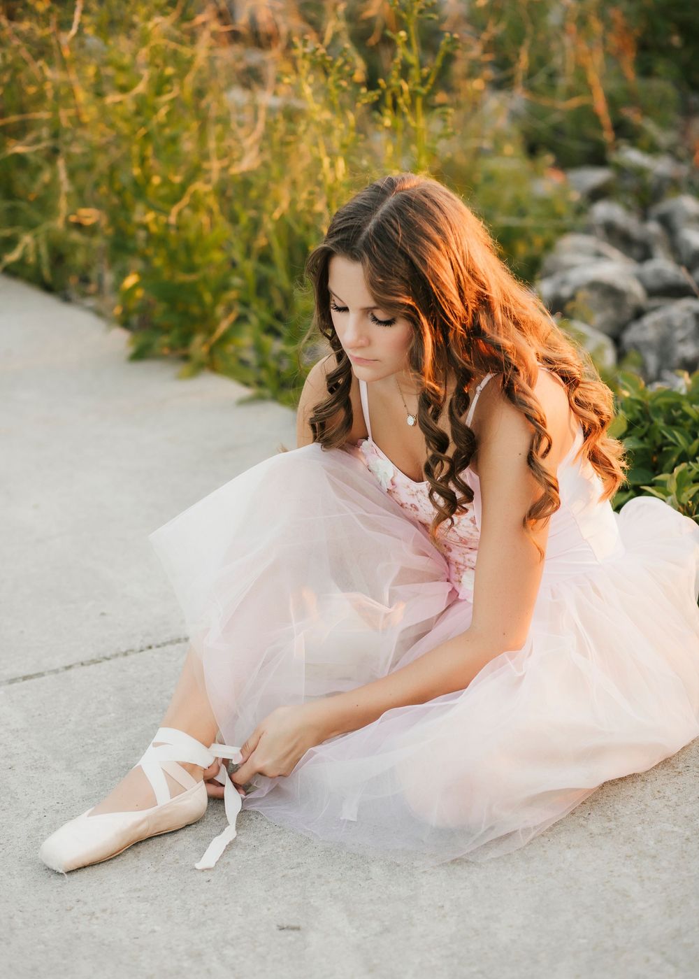 Ballet dancer adjusting pointe shoes while wearing a pink tulle dress outdoors at sunset.