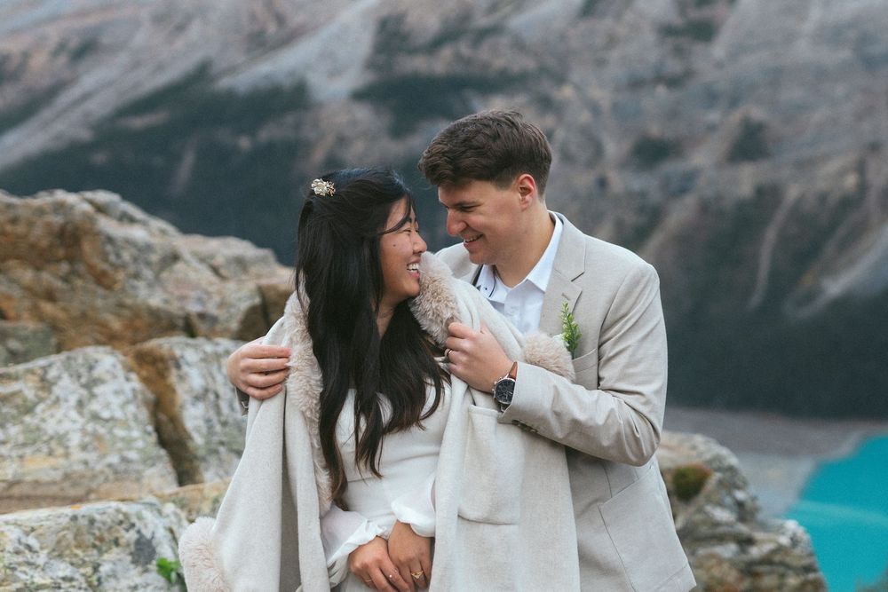 bride and groom laugh in the mountains while he helps her into her coat