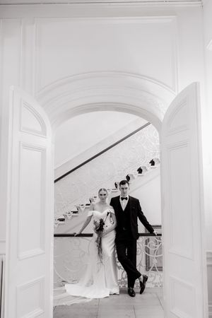A couple stands in an elegant white archway with a curved staircase behind them in black and white photography.