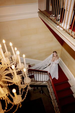 An elegant white chandelier hangs above a winding staircase with red carpeting and black metal railings.