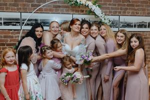 A wedding party poses together against a brick wall with floral decorations and pink bridesmaid dresses.