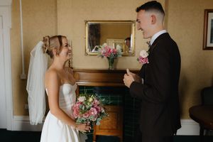 A wedding couple stands in an elegant room during their first look moment with pink floral bouquet details.