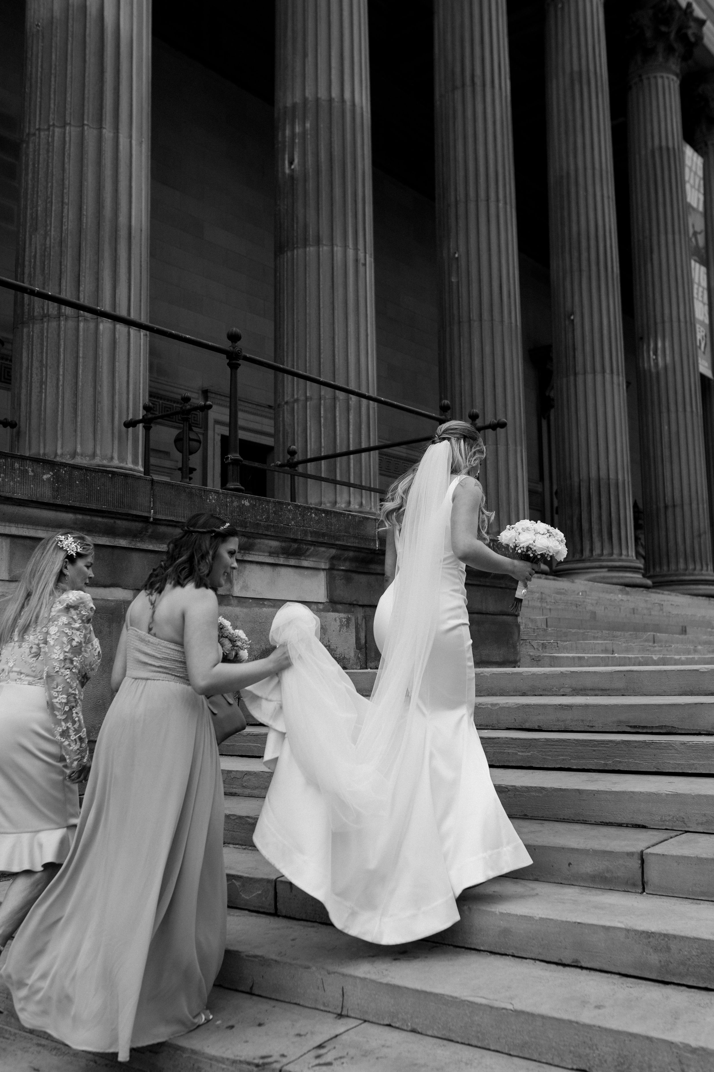 A bride in white dress climbing stone steps with bridesmaids in light gowns helping with the train at a columned building.