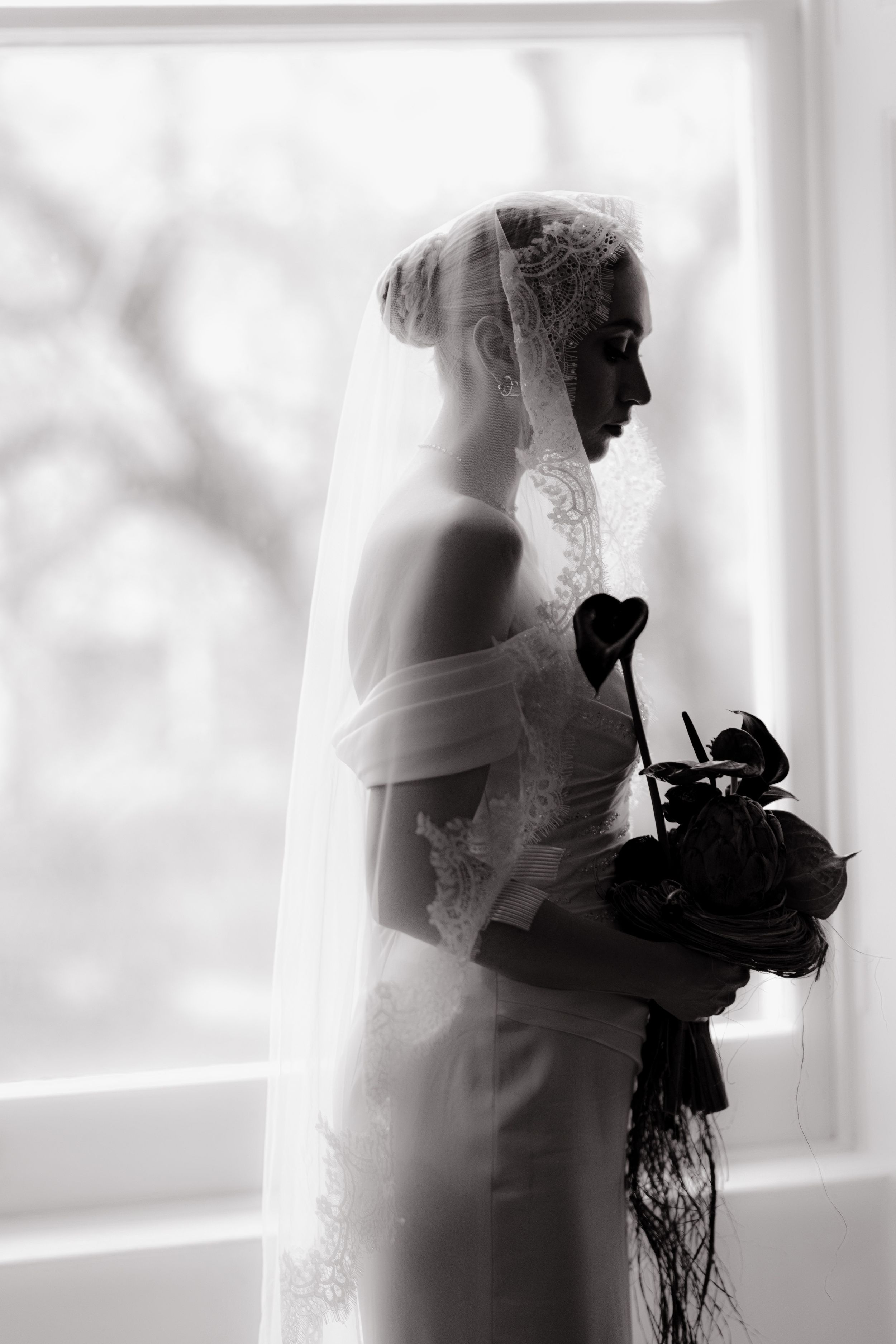 A dreamy black and white silhouette of a bride holding a bouquet near a bright window.