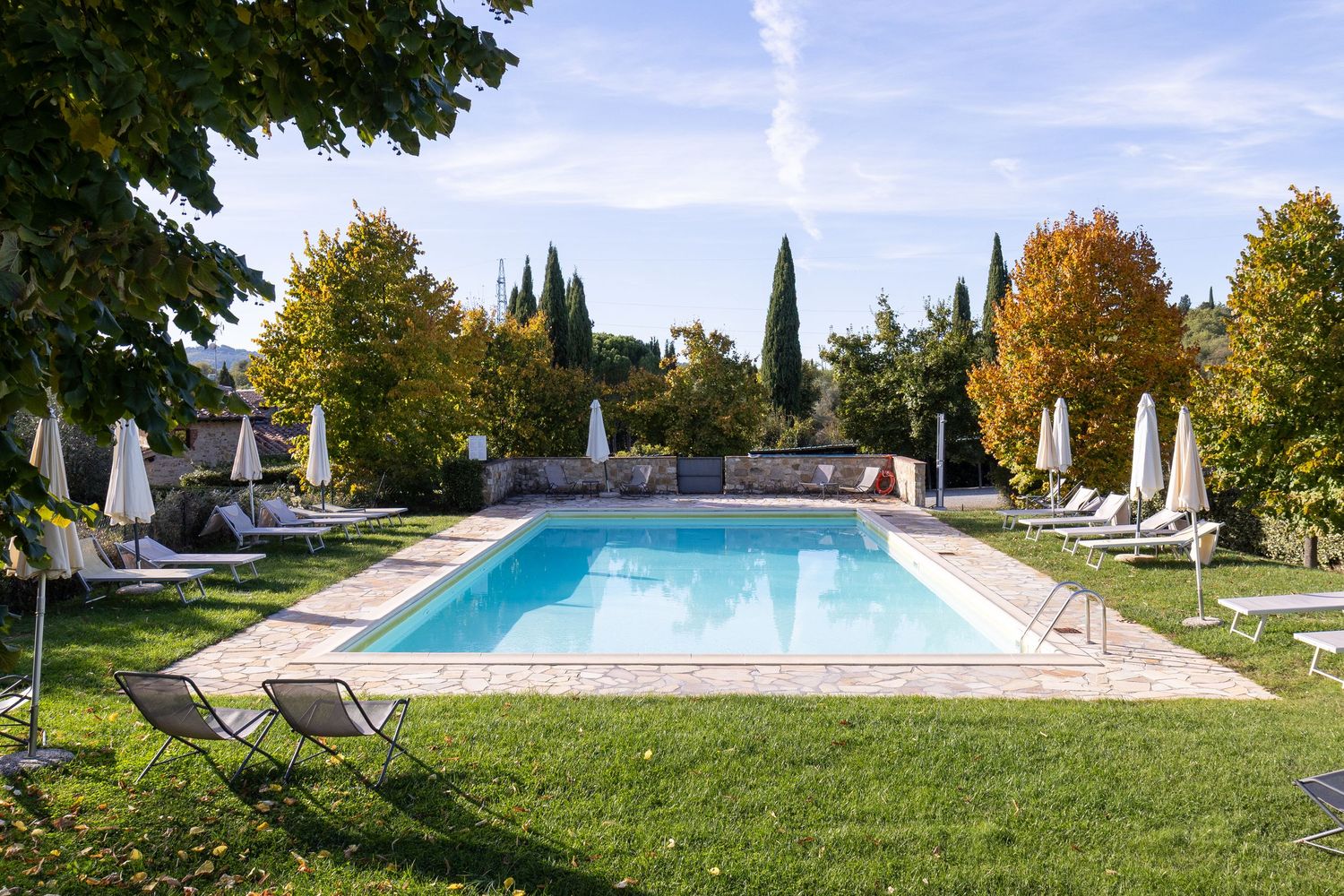 Tuscany property photographer. Luxurious swimming pool surrounded by white lounge chairs and cypress trees in a manicured garden setting.