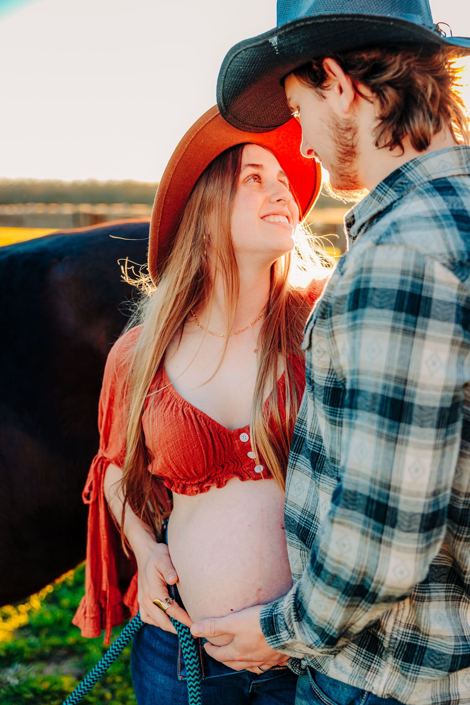 A romantic maternity photoshoot at sunset shows a couple embracing in a plaid shirt and red dress.
