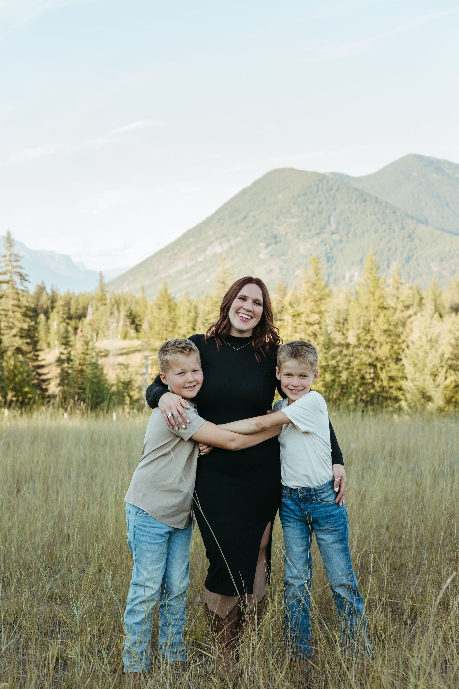Family portrait in a grassy meadow with evergreen trees and mountains in the background during golden hour.