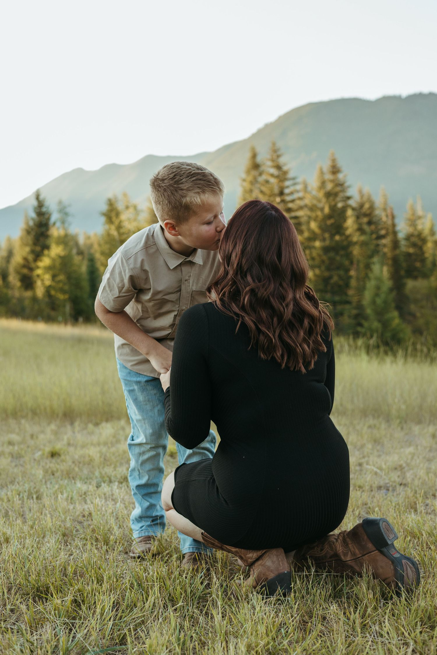 A parent and child sit together in a grassy meadow with evergreen trees and mountains in the background at sunset.