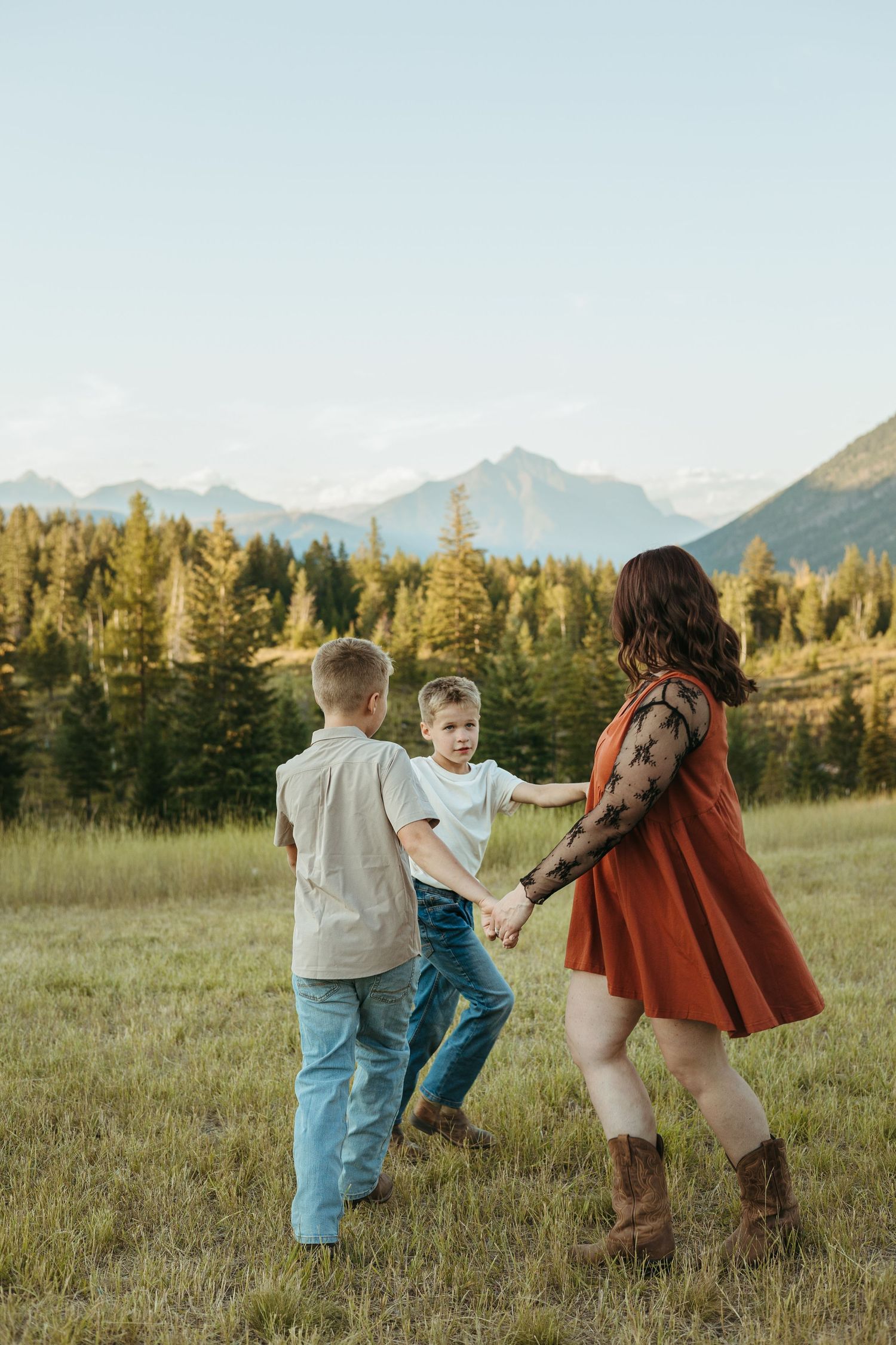 Family plays together in a grassy meadow with mountain views and evergreen trees in the background during golden hour.