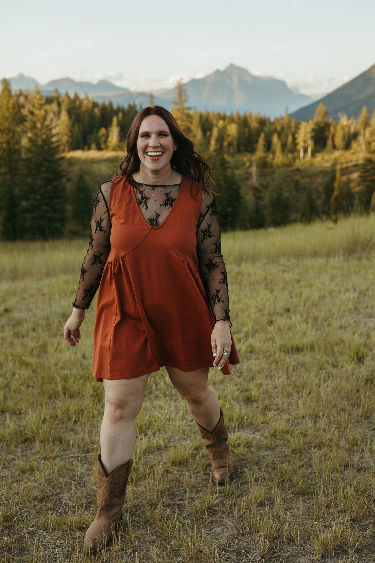A person in a rust-colored top and lace sleeves poses playfully in a mountain meadow at sunset.