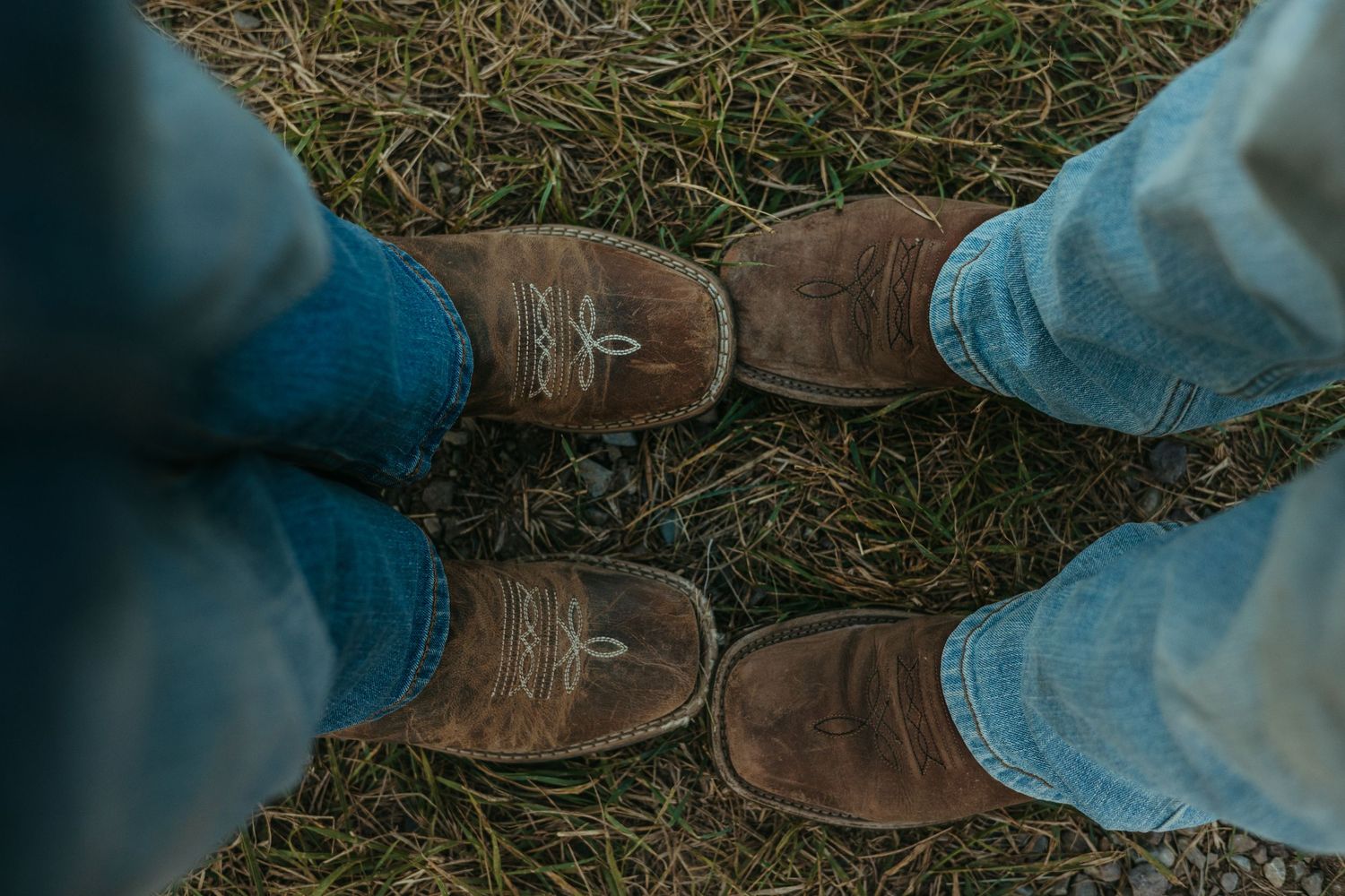 A pair of brown leather boots stands on grass next to light-colored pants legs.