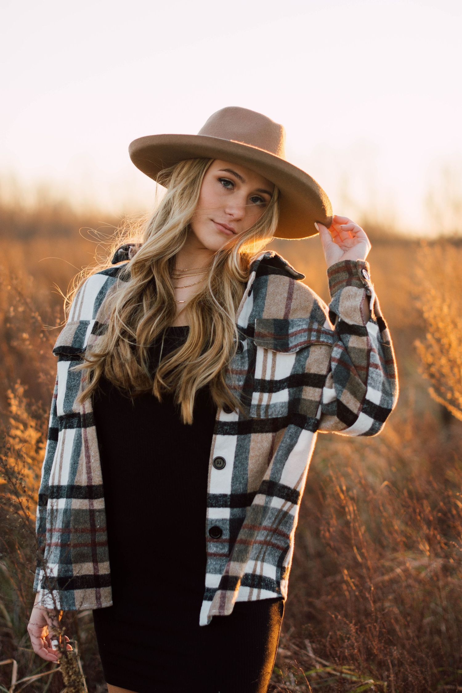 Person in plaid shirt and brown hat poses in golden field at sunset.