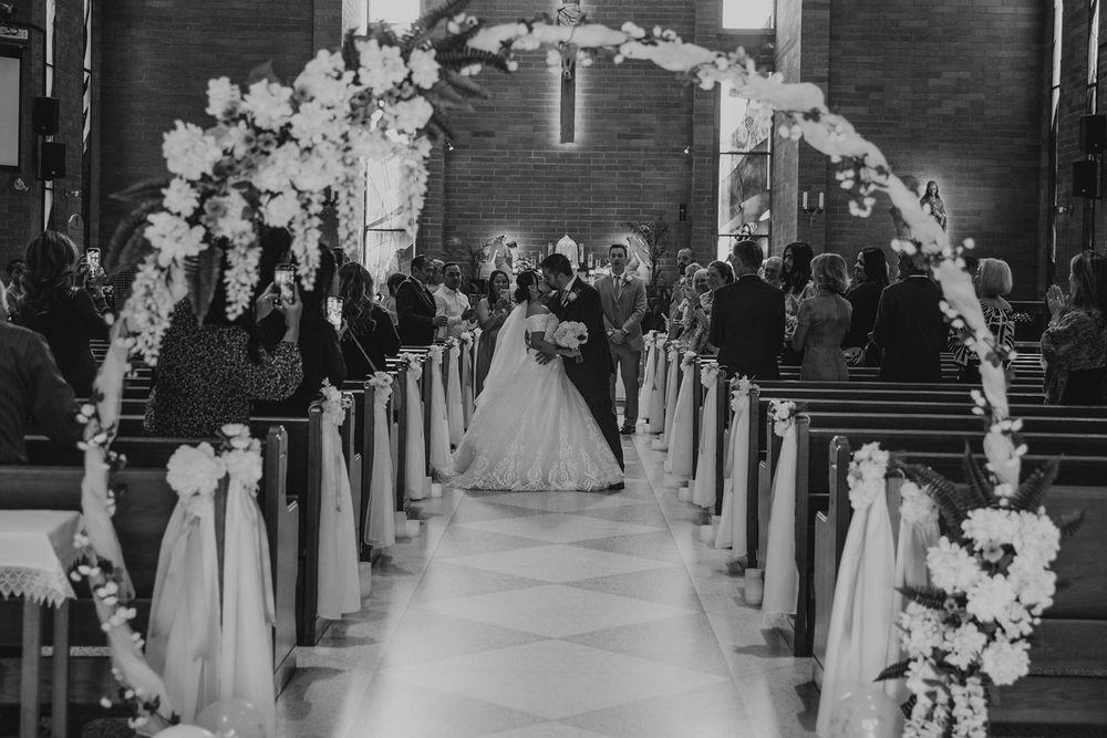 Black and white photo of bride and groom walking down flower-adorned church aisle after ceremony.
