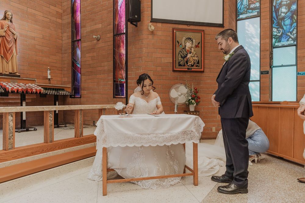 Bride signing wedding documents at altar table while groom stands watching in elegant church ceremony.
