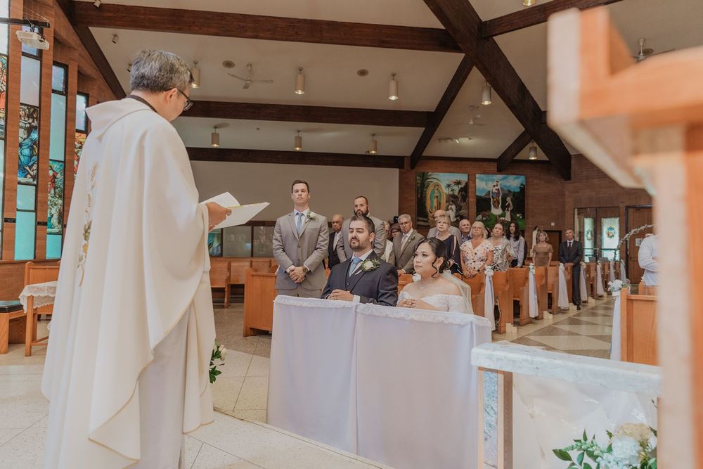 Priest officiating wedding ceremony as bride and groom kneel before congregation in wooden church.
