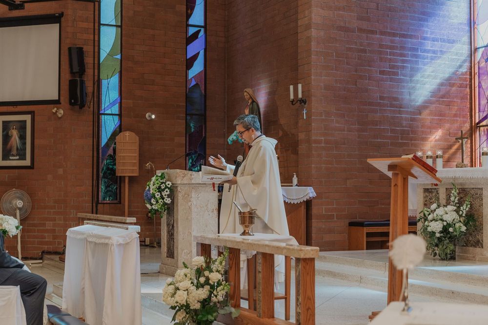 Catholic priest performing wedding Mass at decorated altar with white flowers in brick church.