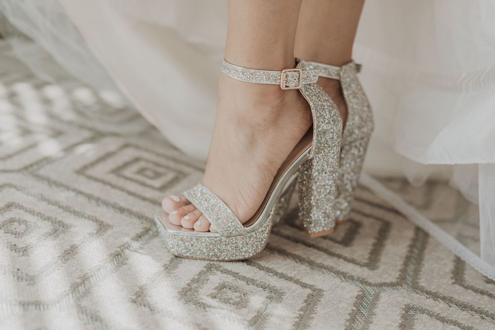 A bride wearing sparkling glitter platform heels with ankle straps stands on a patterned carpet.