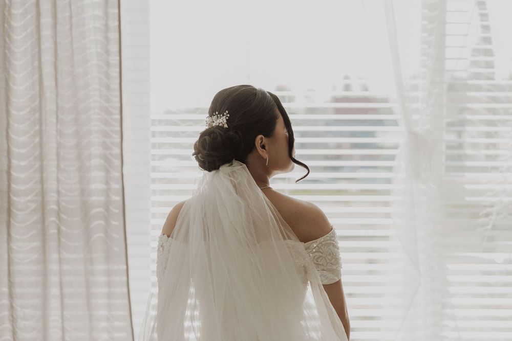 A bride in an off-shoulder wedding gown and veil stands by a sheer curtained window in soft natural light.