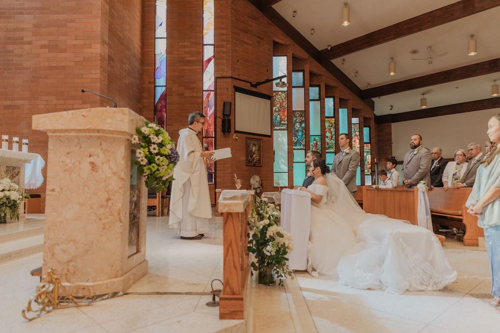 A bride and groom kneel at the altar during their Catholic wedding ceremony in a sunlit brick church.