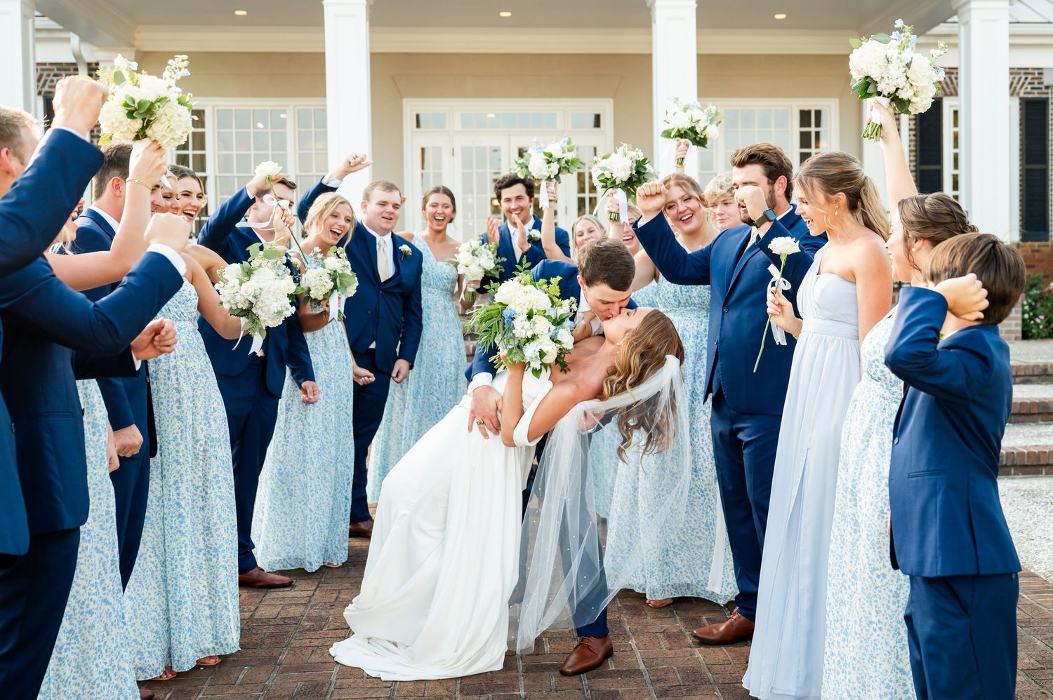 Wedding party in blue dresses and navy suits poses together on a columned porch.