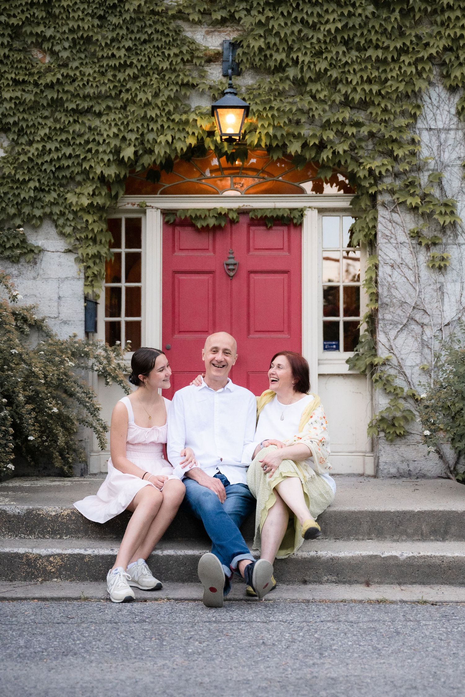 Three people sitting and laughing together on steps in front of a pink door with ivy-covered walls and glowing lantern.