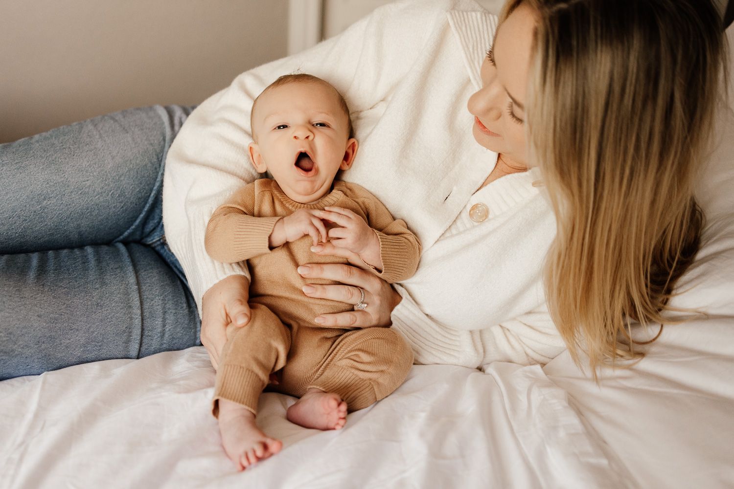 A tender moment between mom and yawning baby lying together on white bedding in natural light.