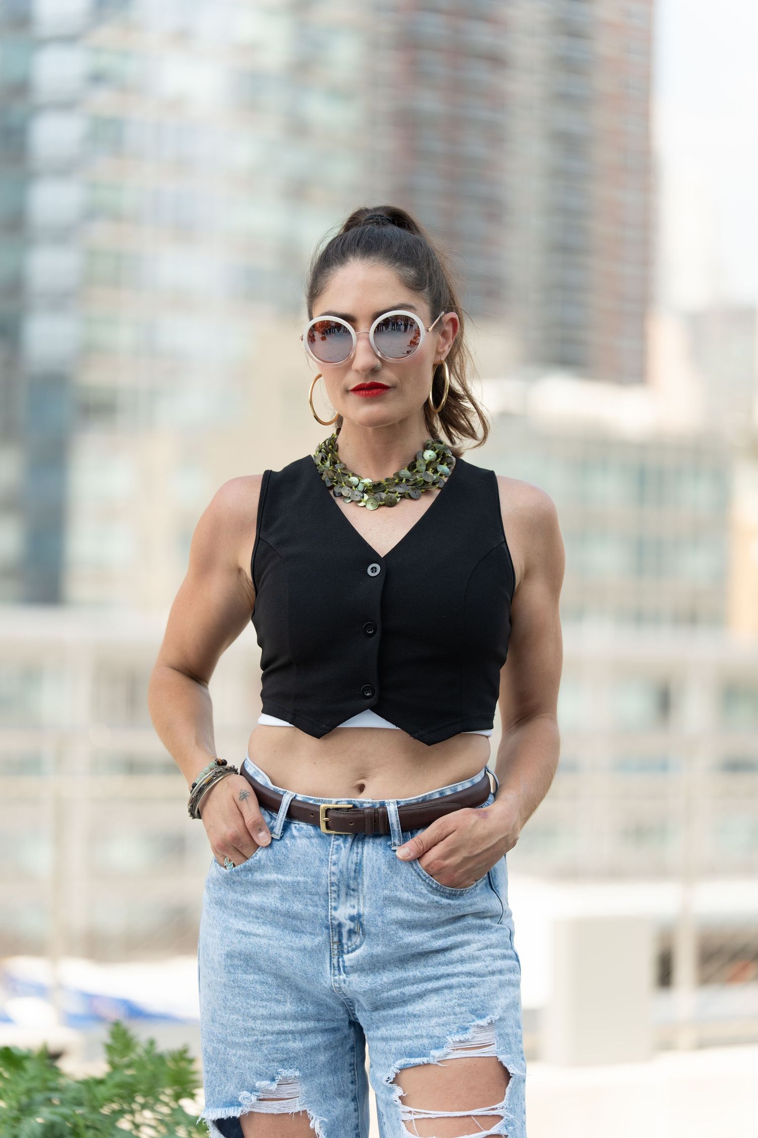 Fashionable woman in cropped black vest and ripped jeans poses outdoors with sunglasses and statement necklace.