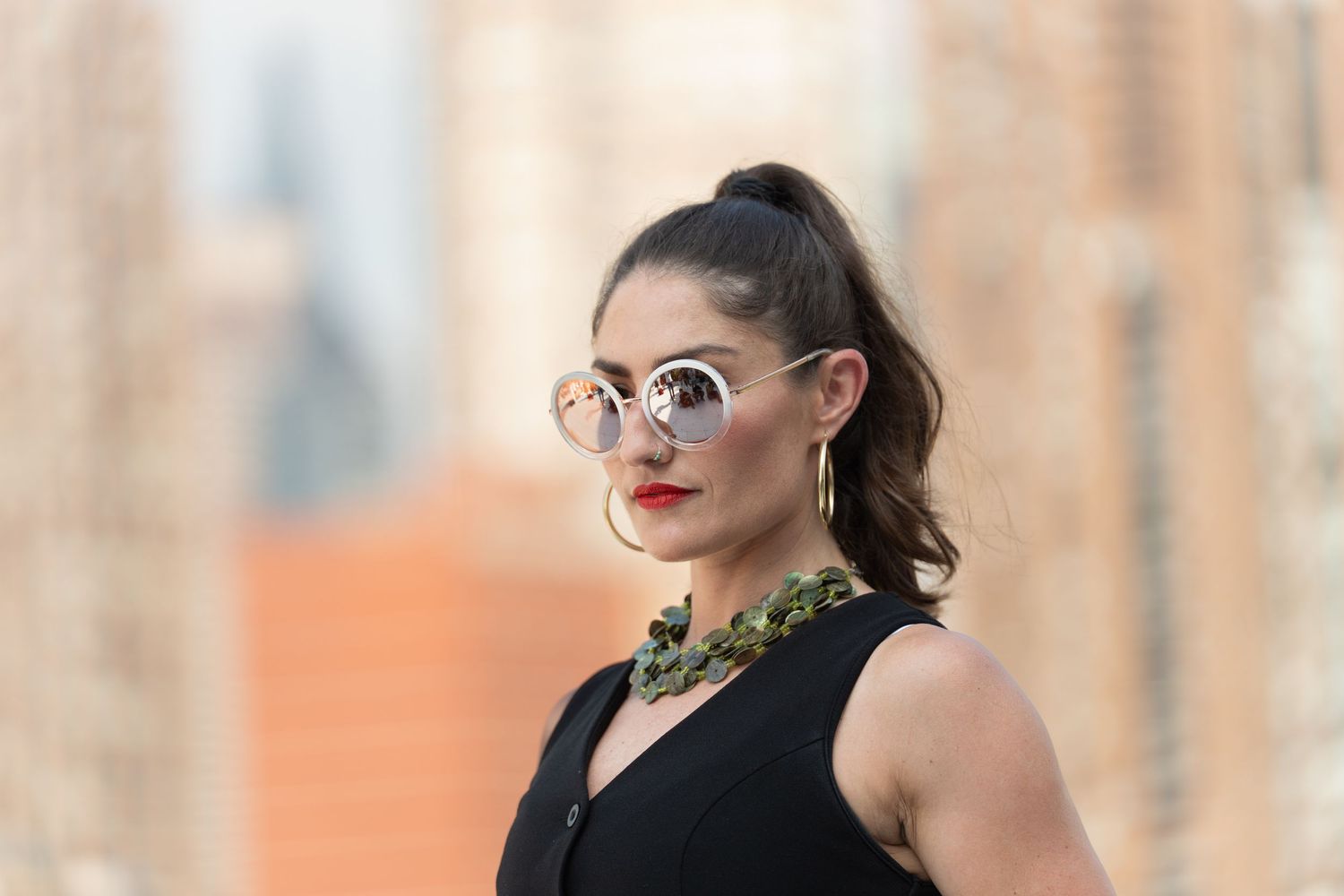 A person in a black sleeveless top with glasses poses against a blurred background.