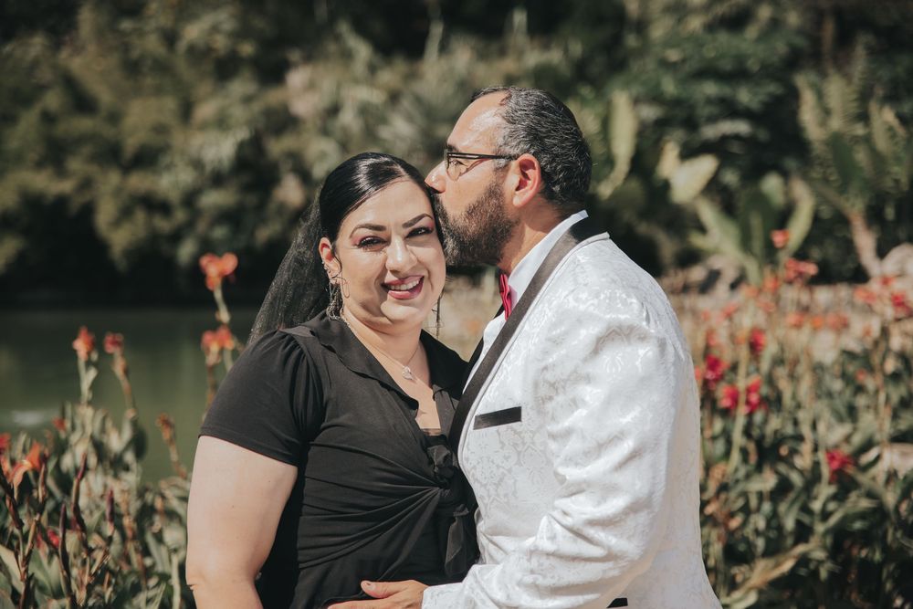 A couple shares an intimate moment in a garden filled with vibrant red flowers on a sunny day.