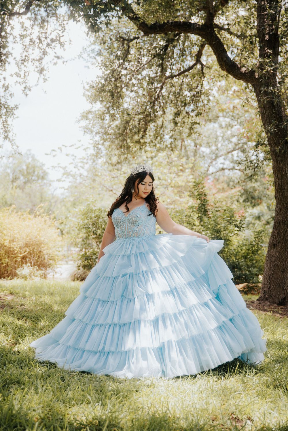 A flowing light blue tiered ball gown twirls gracefully in a sunlit garden setting with trees in the background.