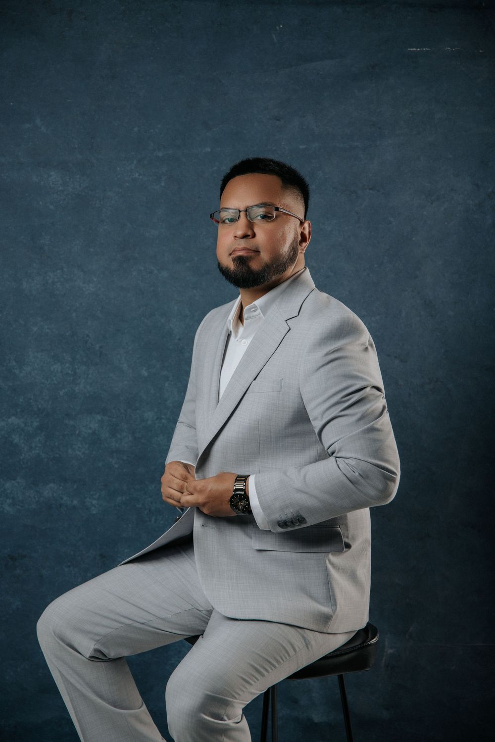 Professional portrait of a person in a light gray suit sitting on a stool against a dark textured backdrop.