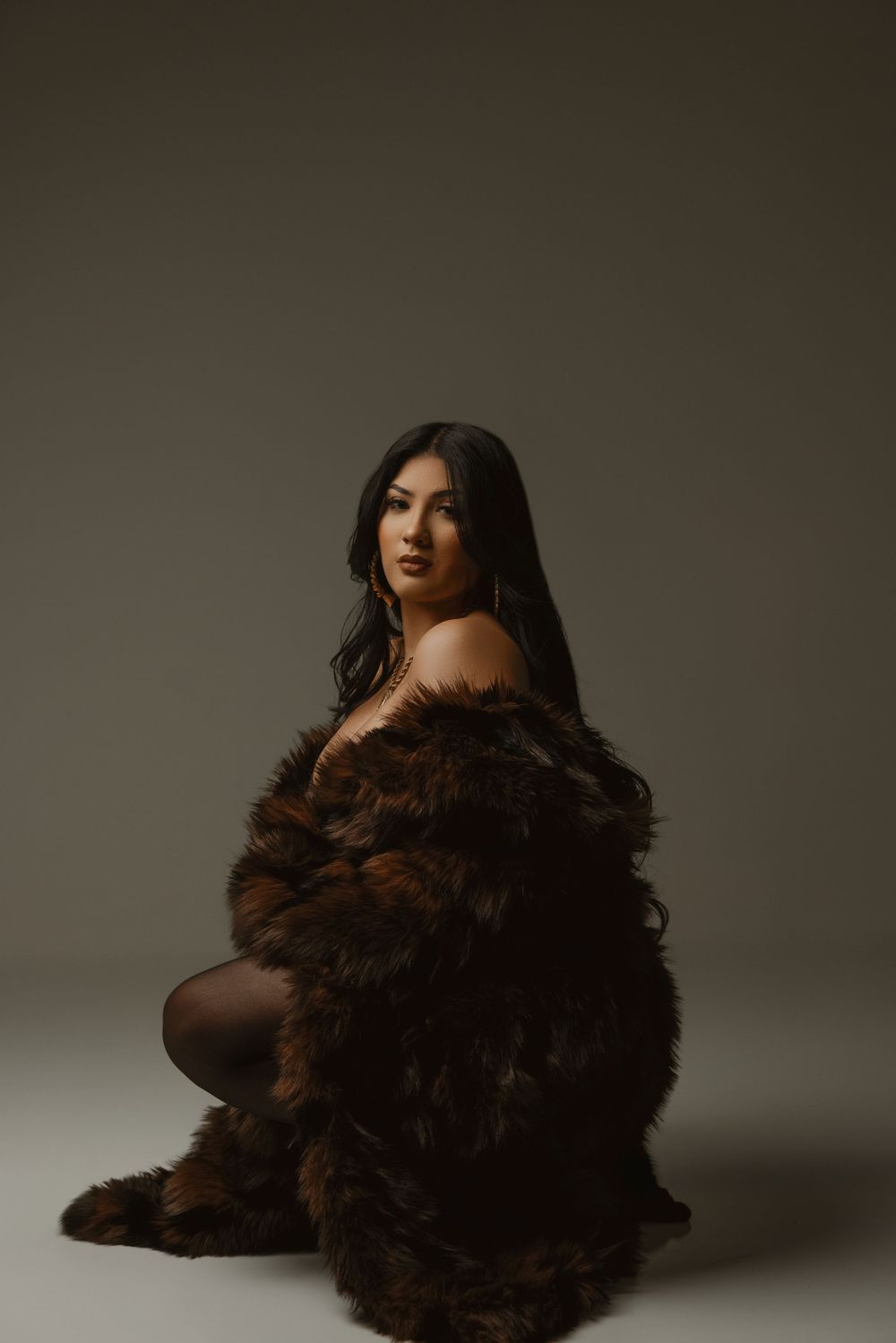 A figure in a dark sheer top poses with dramatic brown fur against a neutral studio backdrop.