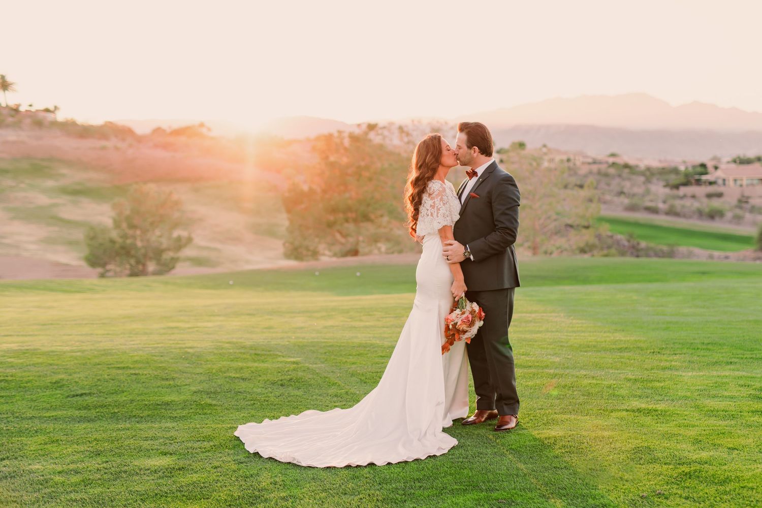 A couple in formal wedding attire walks together through a lush garden path surrounded by trees.
