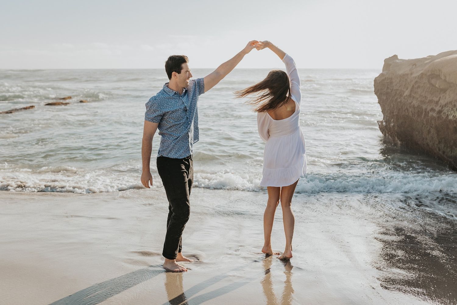 Couple walking and dancing together on a scenic beach during sunset with waves crashing nearby.