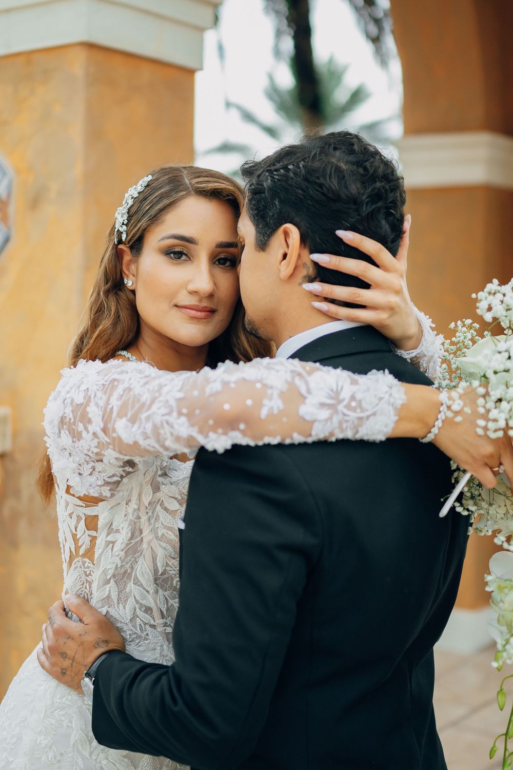 A romantic wedding portrait shows a couple embracing, with the bride wearing a long-sleeved lace gown and holding baby's breath flowers.
