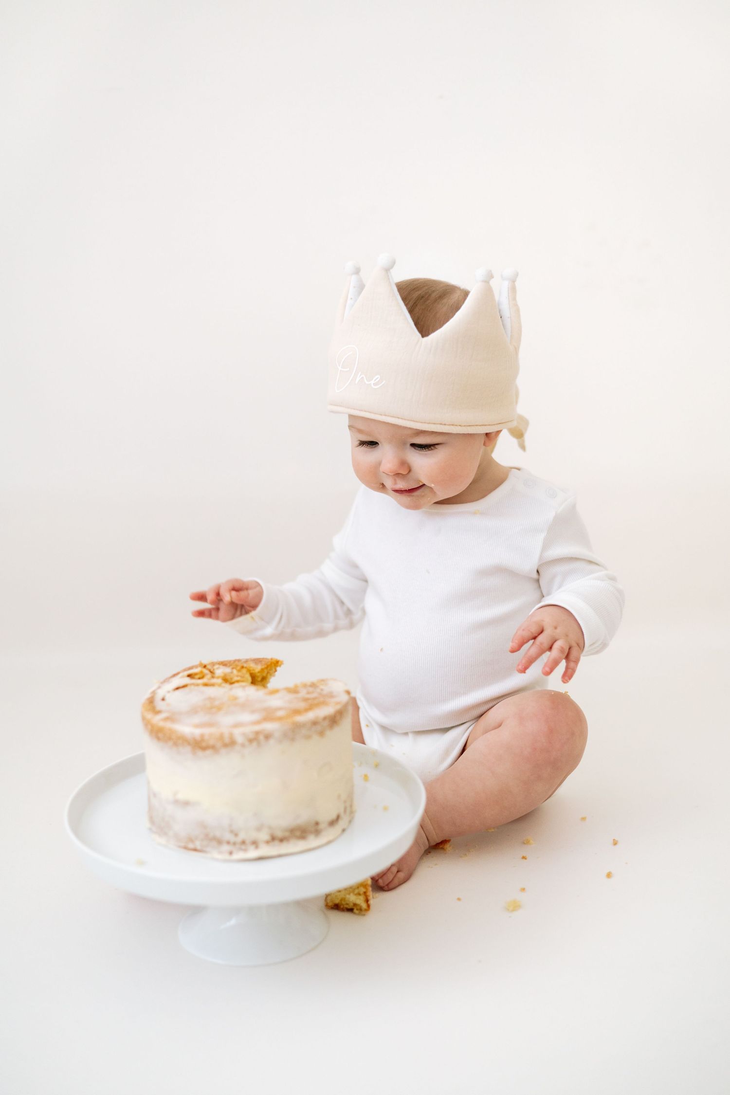 Baby in white onesie reaches for a white frosted cake on plate during cake smash photo session during Birthday shoot based in Nottingham