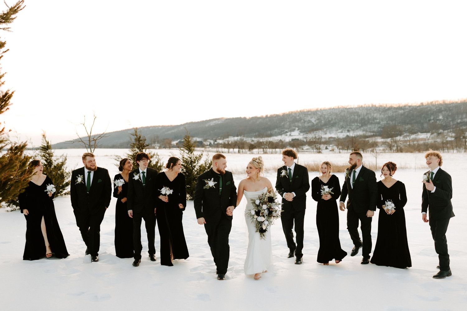 Wedding party in black attire walking through snowy landscape with bride in white gown and groom in black suit.