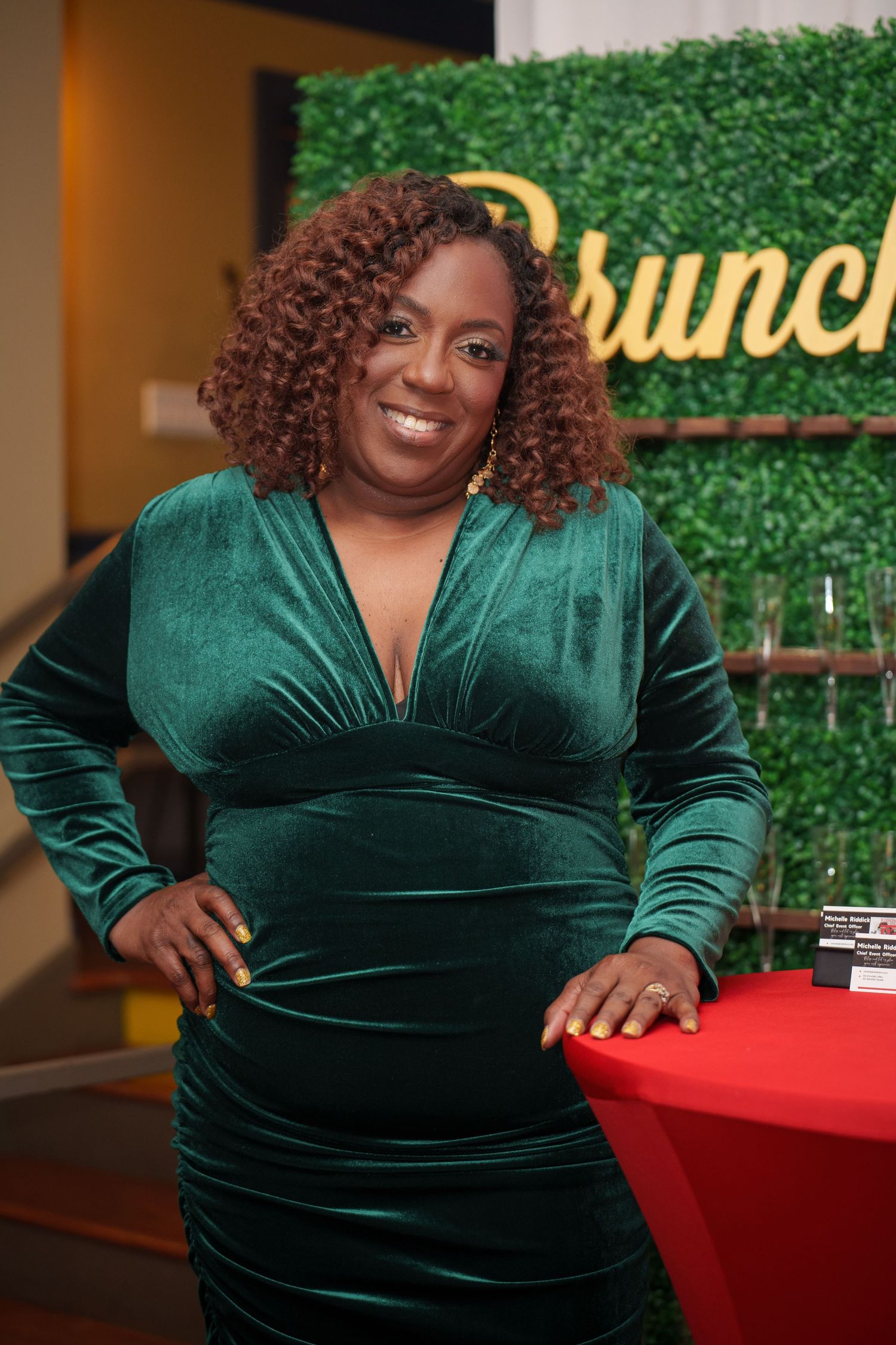 A person in an emerald green velvet dress poses against a decorative green wall backdrop.