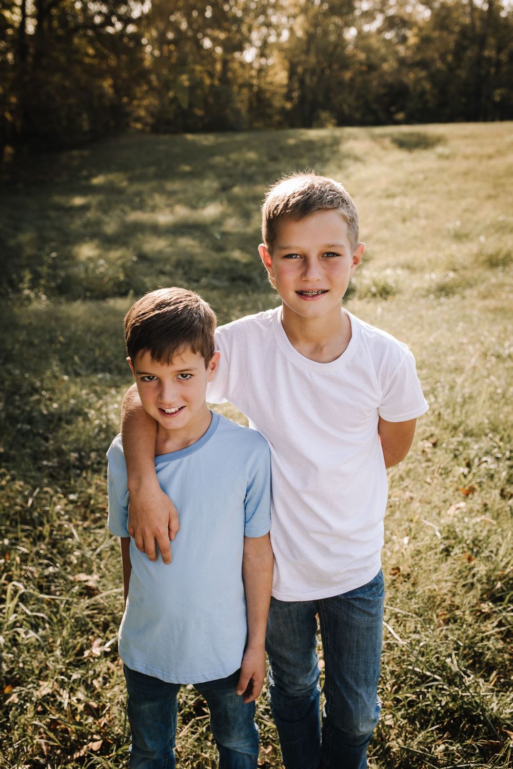 Two children in casual t-shirts and jeans stand together in a sunny field during golden hour.