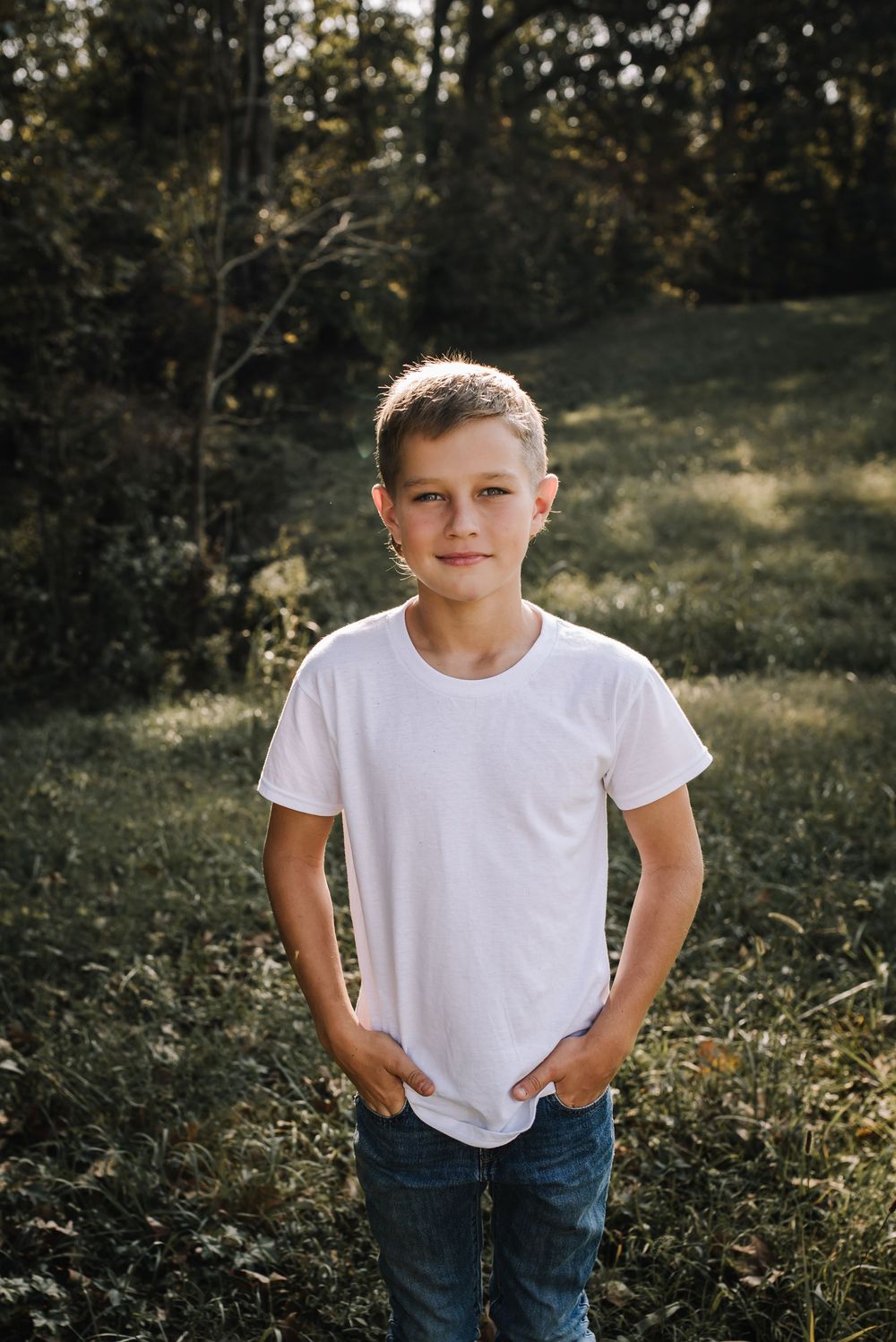 Child wearing white t-shirt and jeans stands outdoors in natural lighting with trees and foliage in background.
