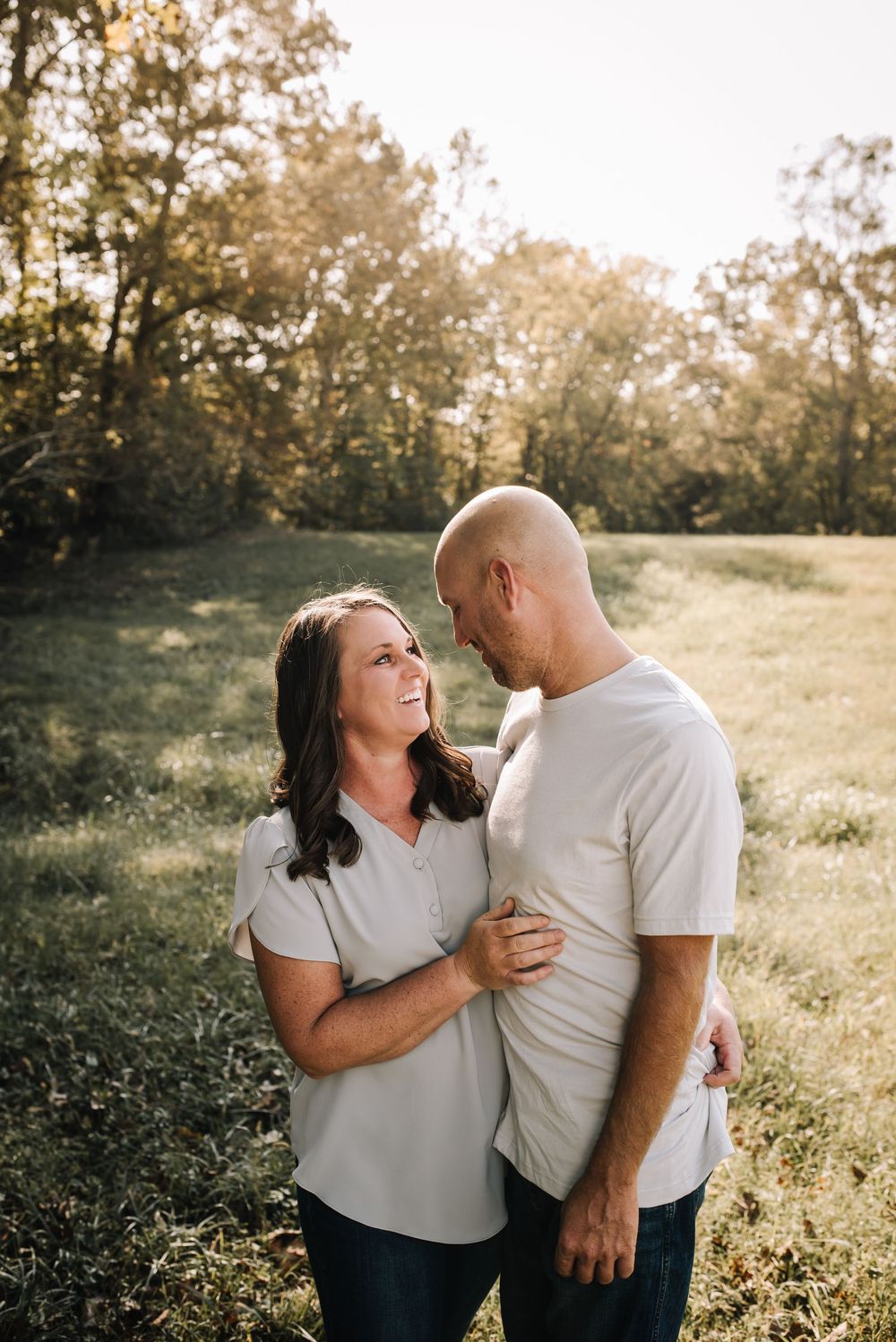 A couple embraces in a sunlit meadow with trees in the background during golden hour.