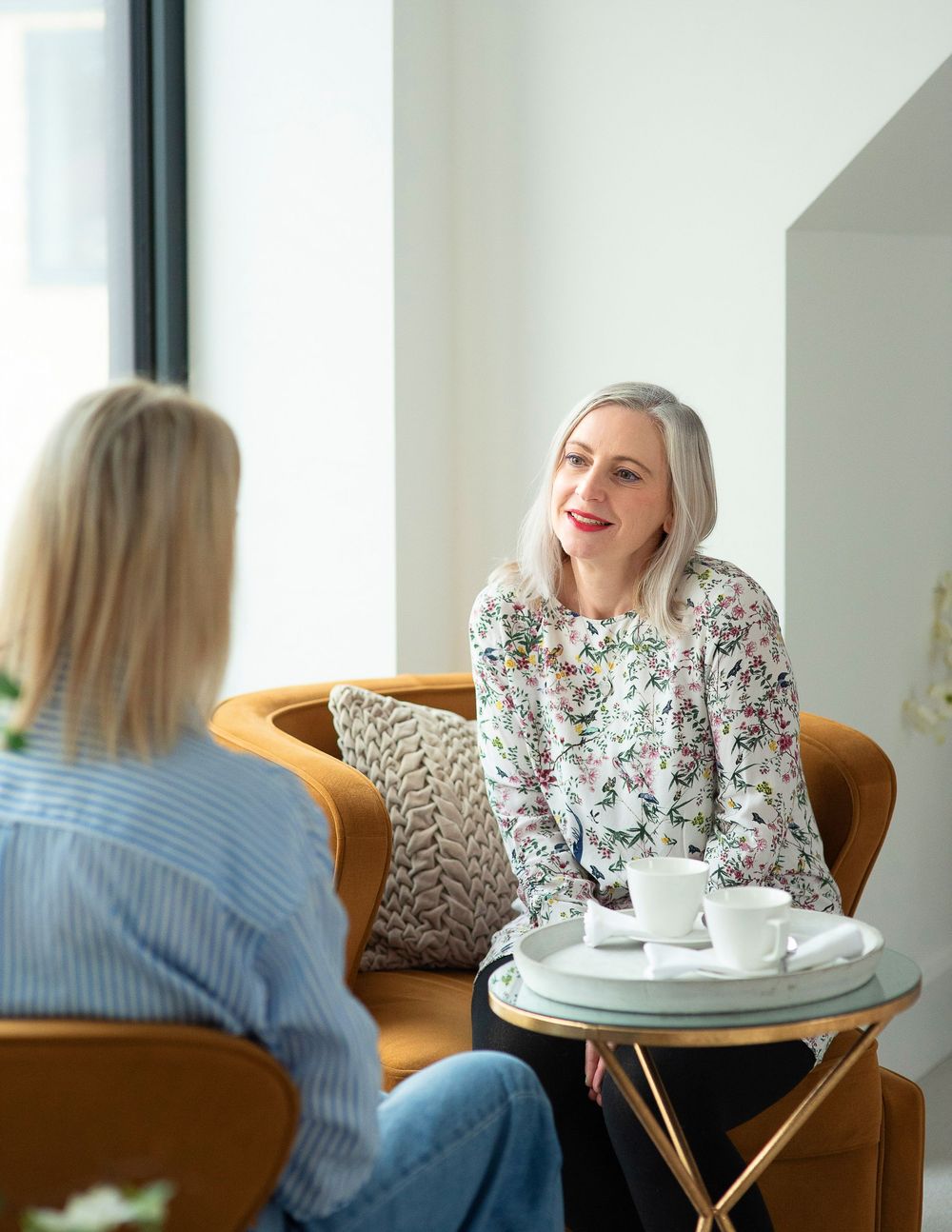Two women sitting at a cafe table with coffee cups having an animated conversation in a bright modern setting.