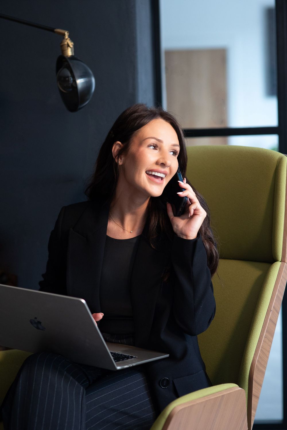 Professional in a black outfit sits at a desk with laptop while laughing and looking away in a modern office setting.